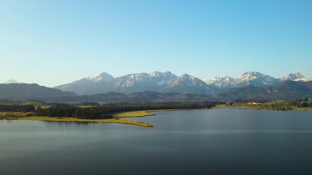 Snow-covered Bavarian Alps rising behind a calm alpine lake near F&uuml;ssen, Germany, with forested foothills under a clear blue winter sky.