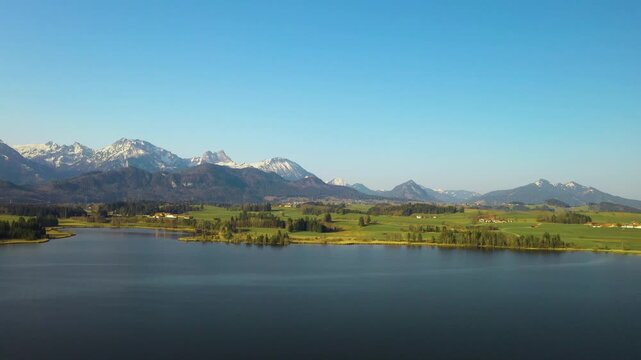 Snow-covered Bavarian Alps rising behind a calm alpine lake near F&uuml;ssen, Germany, with forested foothills under a clear blue winter sky.