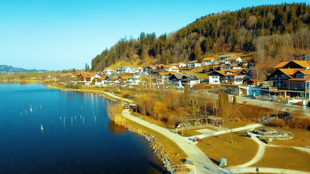 Scenic lakeside village near F&uuml;ssen, Bavaria, Germany, with traditional Alpine houses, forested hills and calm blue water on a clear winter day.