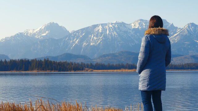 Woman admiring snow-covered Bavarian Alps beside a peaceful alpine lake near F&uuml;ssen, Germany. Scenic winter travel and nature landscape in Europe.
