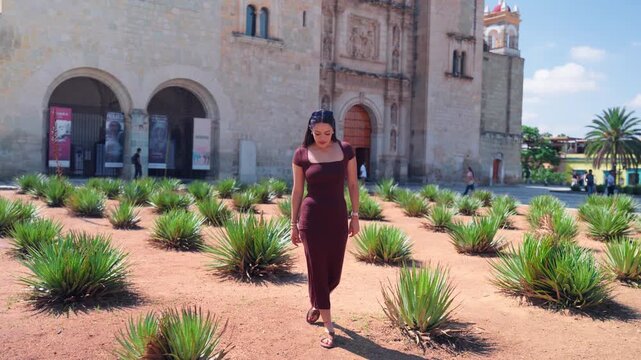 Beautiful Hispanic woman in an elegant dress walking toward the camera and smiling while visiting a famous landmark in Oaxaca, Mexico, on a sunny day during her summer vacation