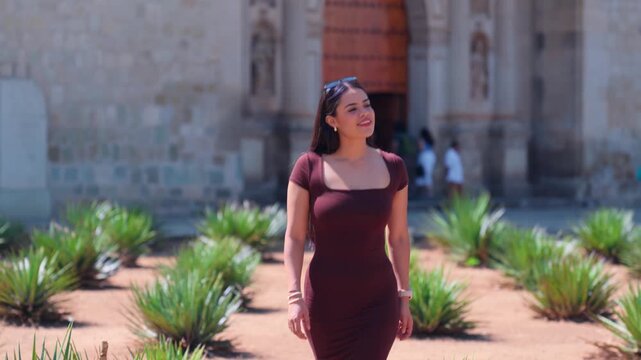 Smiling hispanic woman visiting the historic center of Oaxaca in Mexico, walking through a beautiful garden with agave plants and a colonial church in the background on a sunny day