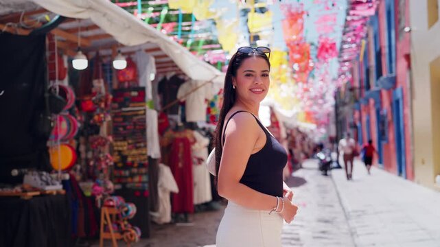 Beautiful Latina woman with long black hair looking up at colorful decorations and then turning to smile at the camera while walking on a traditional street market in Oaxaca, Mexico