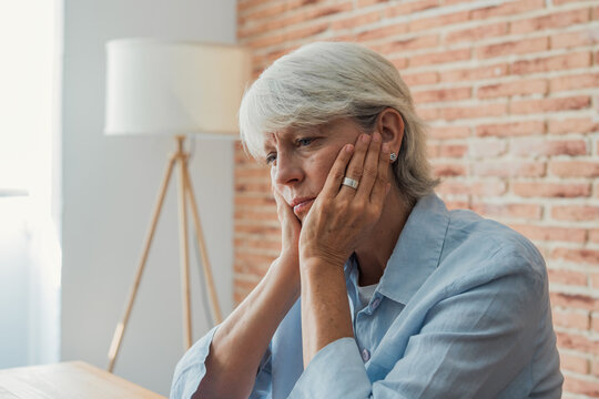 Senior woman experiencing feelings of sadness, loneliness, and depression, sitting indoors with her hands touching her face, contemplating her thoughts in a moment of quiet reflection