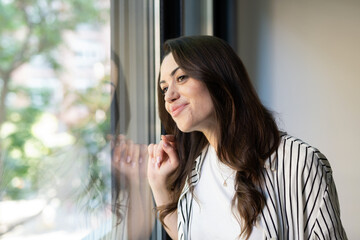 Fototapeta premium Young woman with long brown hair standing by a sunlit window, smiling and daydreaming as she enjoys a quiet, contented moment of calm reflection at home