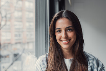 Fototapeta premium Young woman smiling at the camera, standing indoors by a large window with blurred city buildings in the background, exuding confidence and approachability