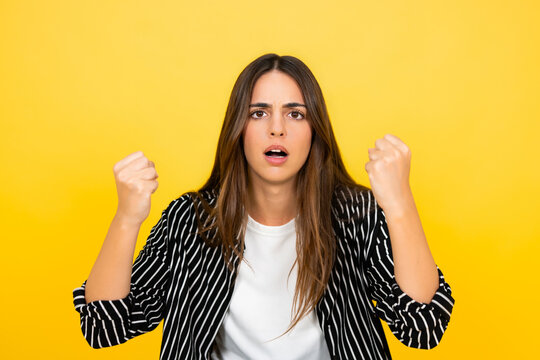 Young woman standing against a yellow background, clenching her fists with a frustrated and angry expression, conveying feelings of outrage, stress, and disappointment