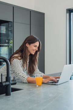 Woman smiling and typing on a laptop computer while leaning on a modern kitchen counter with a glass of orange juice, representing remote work, productivity, and a balanced lifestyle