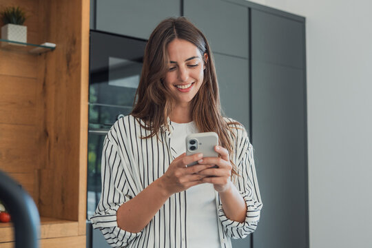 Smiling woman standing in a modern kitchen, holding and using a smartphone, checking messages, browsing social media, or shopping online, representing digital lifestyle and connectivity