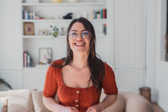 Young woman wearing glasses, dressed in a casual red shirt, looking at the camera and genuinely laughing with happiness and optimism in a bright, modern living room at home