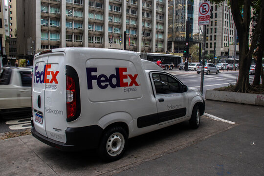 A white FedEx Express Fiat Fiorino cargo delivery van parked on the side of the bustling Paulista Avenue, the main financial heart and historical landmark of Sao Paulo city,