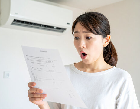shocked japanese woman looking at high utility bill or electricity invoice under air conditioner