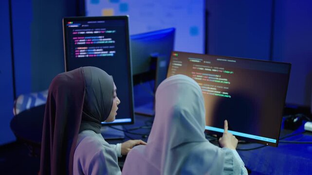 Two diverse Muslim women in hijabs collaborating on software code late at night in a dark blue lit tech office. Female programmers discussing a complex IT project on dual computer monitors.