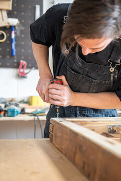 Focused master using electric cordless drill instrument and fastens metal screw in corner of vintage frame, professional carpenter finishing renovation of antique window frame in restoration workshop.