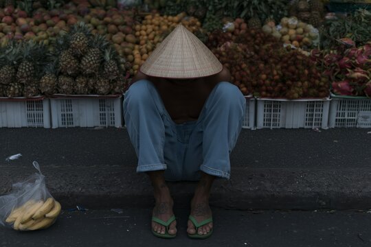 A seated street vendor wearing a conical hat at a vibrant fruit market, surrounded by colorful tropical fruits.