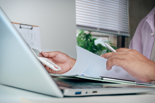 Business professional holding calculator and flipping paper document report on office desk with laptop computer setup, concept of financial calculation, accounting analysis, and corporate paperwork.