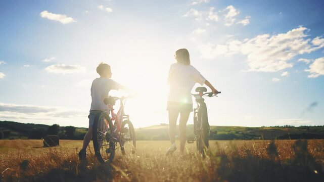 Two joyful children riding bikes along rustic country path with golden grasses. Happy summertime in countryside, back view of teens strolling bicycles and walking in beautiful farmland in sunny day