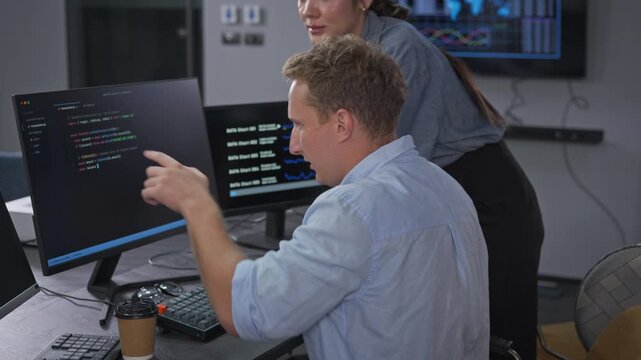 Focused developer types code on a dual monitor workstation while a colleague stands behind him, working through a software build in a dark modern technology office.
