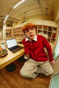 Portrait of Caucasian man looking at camera while sitting at desk with laptop in modern library. Image ideal for youth education, remote learning, and digital lifestyle campaigns, vertical