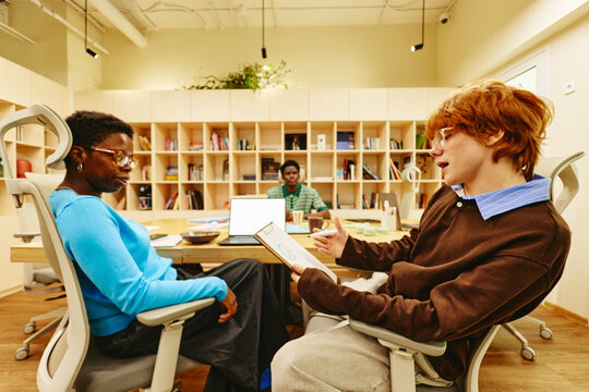 Young adult Caucasian man holding clipboard and talking to Black woman in modern office. Image represents corporate communication and teamwork, suitable for business marketing