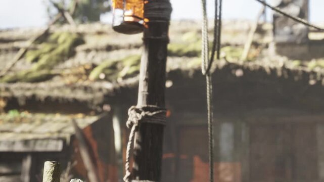 A lantern is hanging on a wooden post. The post is tied with rope. In the background, there is a rustic structure covered in moss. The scene appears to be outdoors in daylight.