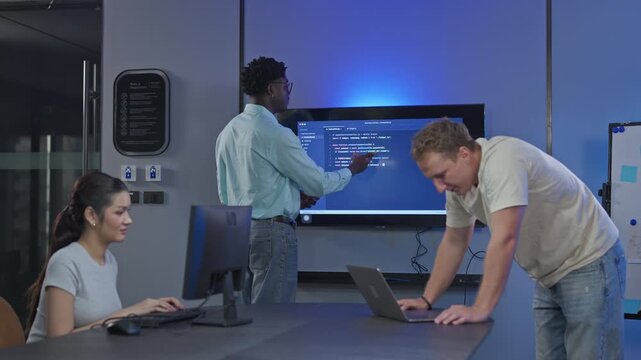 Three developers work through code shown on a large wall display, one pointing at the screen while another types on a laptop during an evening debugging session.