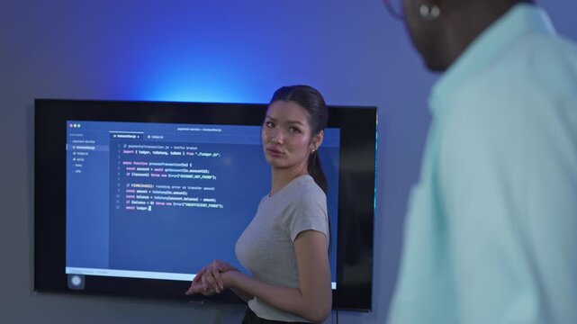 Smiling young female developer gestures toward code displayed on a large wall screen while explaining her implementation to a colleague in a modern blue lit tech office.