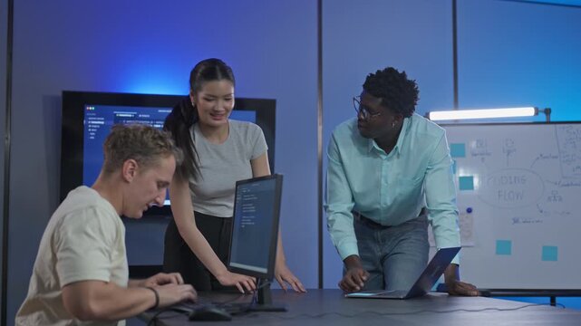 Three diverse software developers smile and talk together around their desks, with code on the wall screen and an agile flow diagram on the whiteboard behind them.