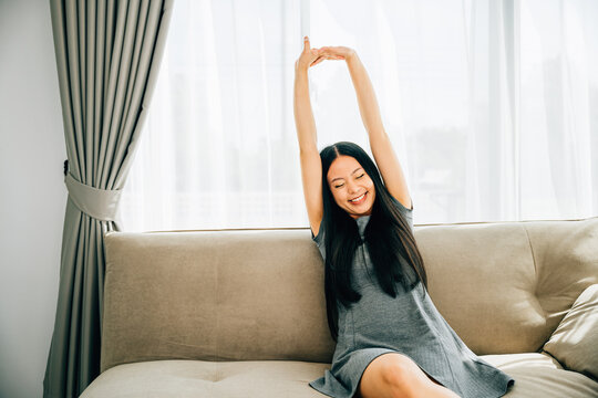 Young woman relaxes on sofa stretches with raised arms after sitting. Lifestyle of a cheerful entrepreneur in a luxury living room. Wellbeing joy and carefree resting at home.