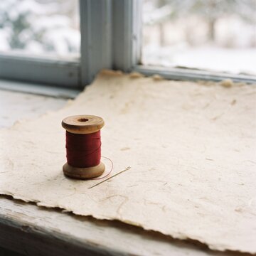 Vintage wooden spool with red thread and sewing needle on handmade textured paper by window, cozy handicraft craft concept.