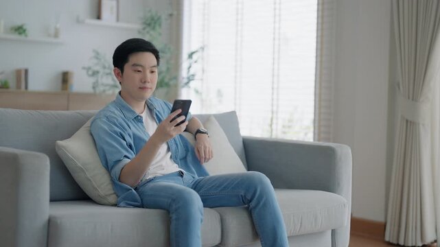 Smiling handsome young Asian man sitting on cozy couch, using funny mobile apps in living room. man at home, doing online shopping, messaging friends, posting on social media