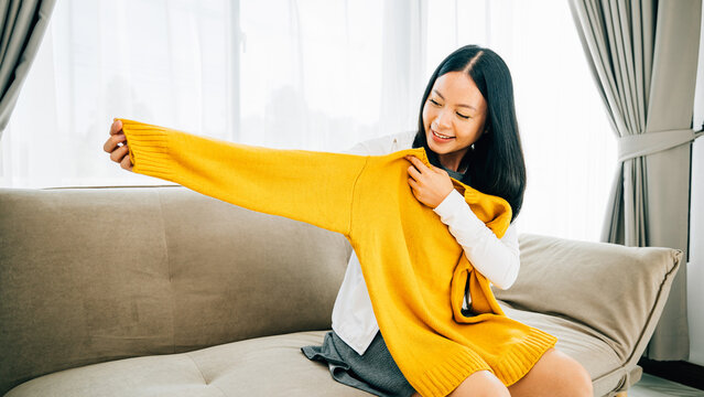 At home an Asian woman happily opens a box with a shirt smiling. Satisfied shopper unboxes online order showcasing new clothes. Delivery and home shopping concept.