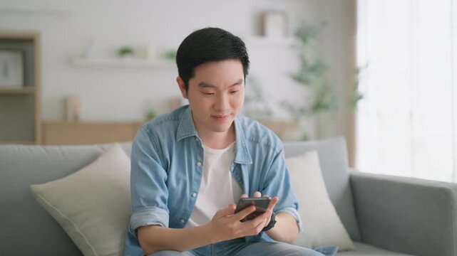 Smiling handsome young Asian man sitting on cozy couch, using funny mobile apps in living room. man at home, doing online shopping, messaging friends, posting on social media
