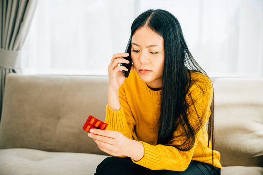 Highlighting home consultation, Asian woman takes medicine consults doctor via phone while sitting on a couch. Depicting healthcare quarantine and social distancing amid coronavirus.