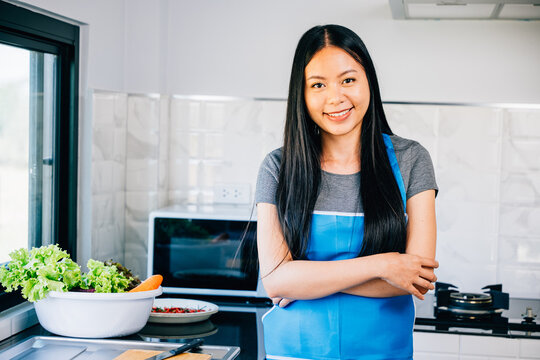 An Asian woman a cheerful housewife stands in a modern kitchen arms folded cooking lunch or dinner. Portrait of a smiling charming woman mixing a delightful meal with joy at home.