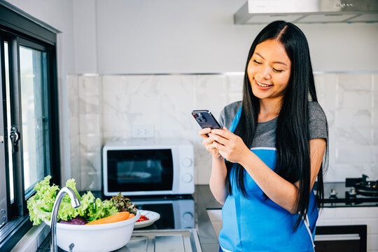 In a cheerful kitchen ambiance a smiling woman uses her cellphone while cooking dinner. Emphasizing multitasking and technology use for a cheerful housewife at home. mobile phone