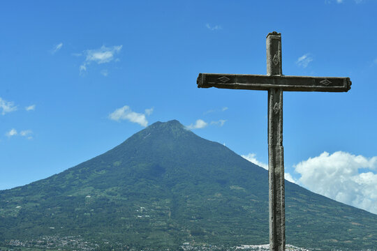 Cruz de piedra con vista hacia el cielo azul y volc&aacute;n de agua en Antigua Guatemala, espacio para texto al lado izquierdo.