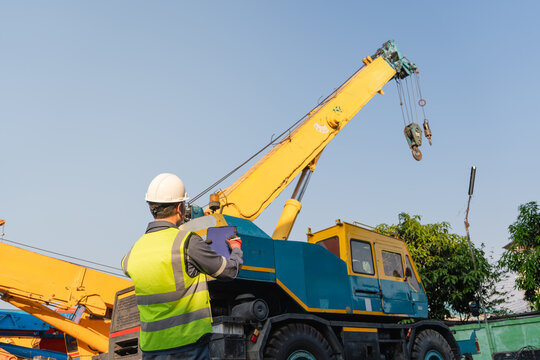 Massive mobile crane undergoes thorough visual inspection as one male engineer wearing a hardhat holds his digital tablet