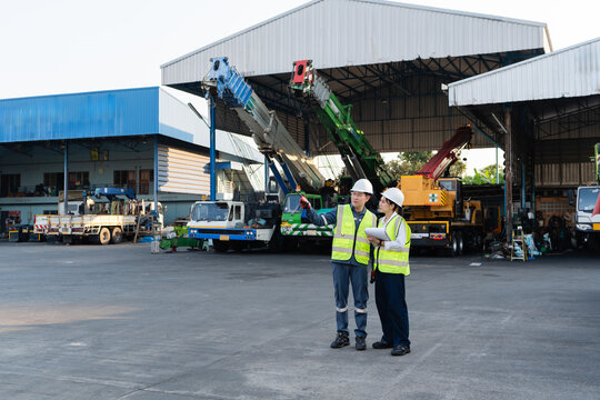 Hydraulic crane booms undergo inspection while male engineer points with radio and female colleague in safety vest holds clipboard