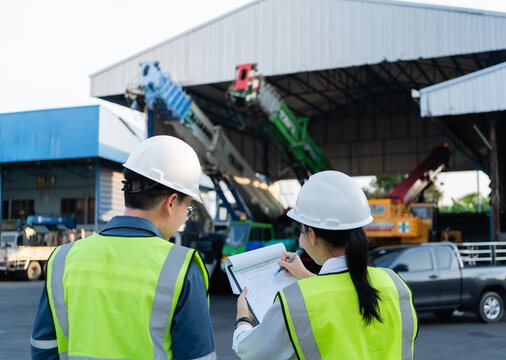 Safety checklist on clipboard for lifting operations is updated by female engineer while male colleague in helmet observes near cranes