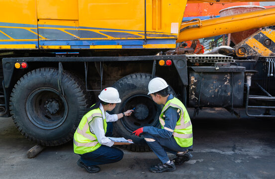 At a lifting site, safety operations involve male and female technicians inspecting a yellow truck crane tire using a clipboard
