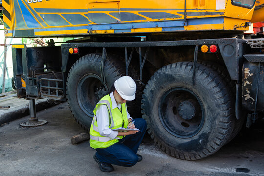 Near the crane outrigger, an inspection proceeds while one female engineer in a hard hat squats writing on a clipboard