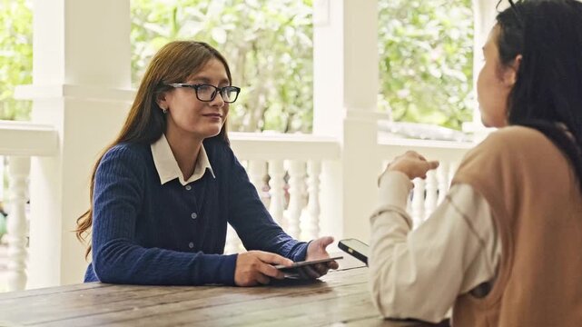 Two women talk at a wooden table on a bright porch, one holding a tablet while listening as the other gestures. A relaxed outdoor meeting with greenery in the background.
