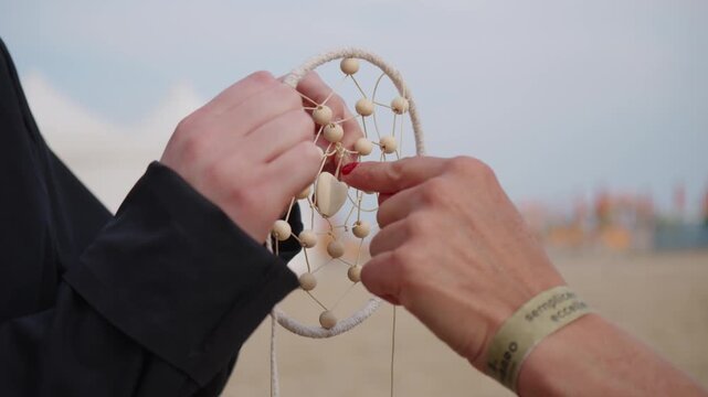 Close-up of hands crafting a handmade dream catcher with wooden beads and woven threads outdoors, showing detailed creative artisan work.