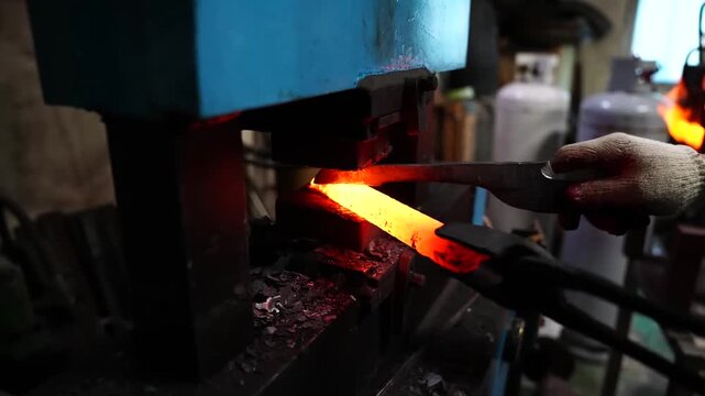 Tapering a glowing steel bar under a power hammer while gas cylinders stand in the workshop background