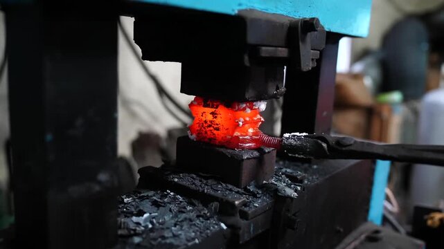 Power hammer compressing a white hot layered billet as scale and sparks fall away inside a knife making shop