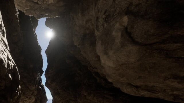 A person stands inside a narrow gap between large rocks in a desert area. The sun shines through the opening above, illuminating the scene. The rocks are rugged and weathered.
