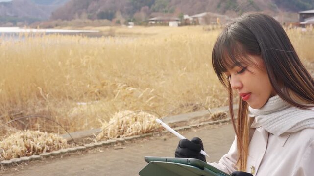 Asian woman drawing on digital tablet by Lake Kawaguchi near Mount Fuji in Japan. Female traveler sketching outdoors during winter vacation, scenic sightseeing, travel lifestyle and Japanese tourism