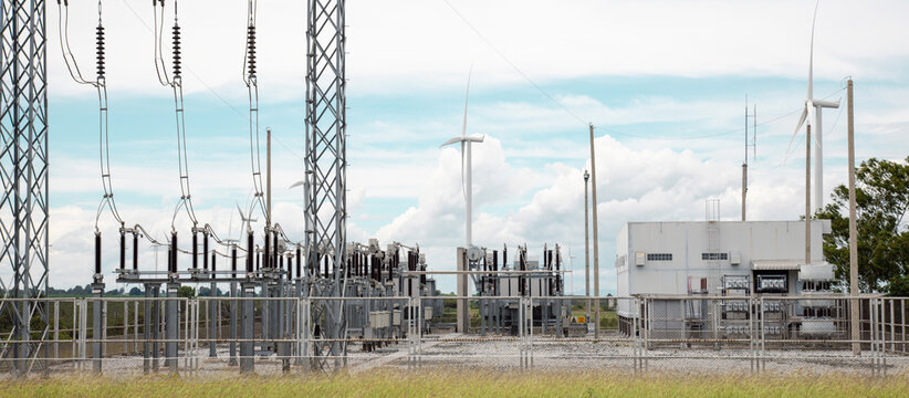 High-voltage electrical substation facility connected to a green wind energy turbine farm under a cloudy sky.