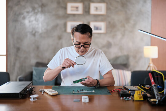 Mature Asian man looks at and considers circuit board details with magnifying glass, focusing on precision checks for home electrical appliance DIY fix, electronics component hobby, handyman workshop.